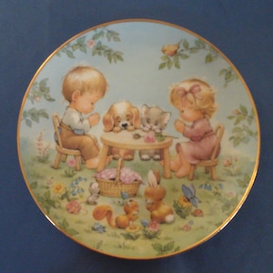 May include: A porcelain plate with a colorful illustration of two children praying with their animal friends. The scene is set in a garden with flowers, trees, and a small table. The plate has a gold rim.