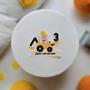 May include: A white cake decorated with a sticker featuring a child in a yellow construction vehicle, the number 3, and the text "Aiden's 3rd Birthday". The cake is surrounded by yellow and orange decorative elements.