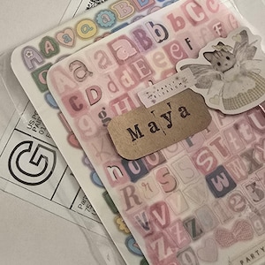 K&ouml;nnte beinhalten: Transparente Aufkleberb&ouml;gen mit Buchstaben in verschiedenen Farben, darunter Rosa, Blau und Gelb. Ein kleines Etikett tr&auml;gt die Aufschrift "Maya". Ein Aufkleber einer Fee ist ebenfalls zu sehen.