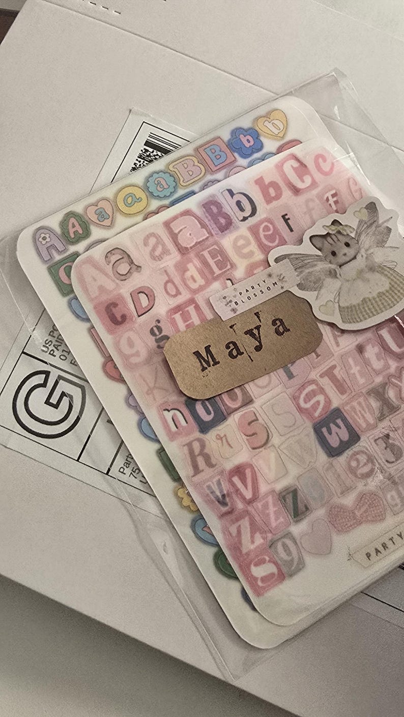 K&ouml;nnte beinhalten: Transparente Aufkleberb&ouml;gen mit Buchstaben in verschiedenen Farben, darunter Rosa, Blau und Gelb. Ein kleines Etikett tr&auml;gt die Aufschrift "Maya". Ein Aufkleber einer Fee ist ebenfalls zu sehen.