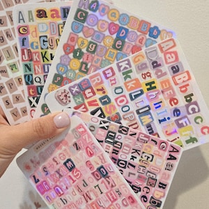 K&ouml;nnte beinhalten: Eine Sammlung von Buchstabenaufklebern in verschiedenen Farben und Schriftarten. Die Aufkleber sind auf transparenten B&ouml;gen angeordnet, einige mit holografischen Effekten. Die Gesamt&auml;sthetik ist farbenfroh und dekorativ.