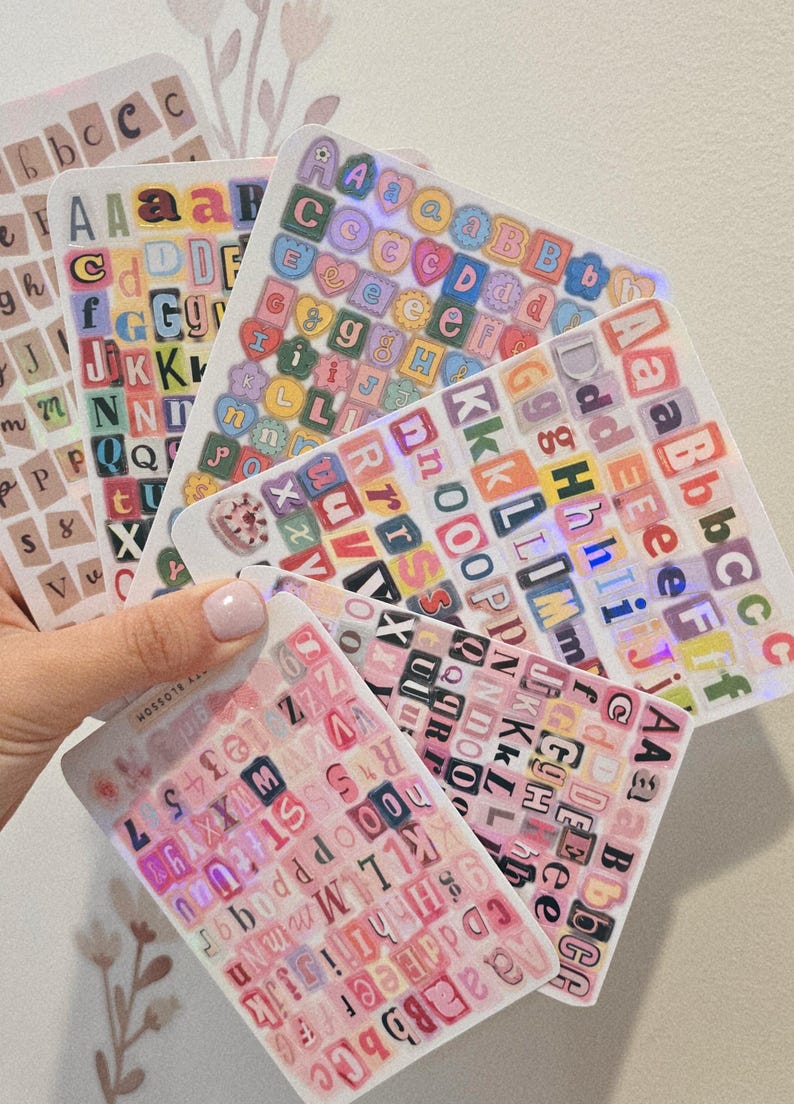 K&ouml;nnte beinhalten: Eine Sammlung von Buchstabenaufklebern in verschiedenen Farben und Schriftarten. Die Aufkleber sind auf transparenten B&ouml;gen angeordnet, einige mit holografischen Effekten. Die Gesamt&auml;sthetik ist farbenfroh und dekorativ.