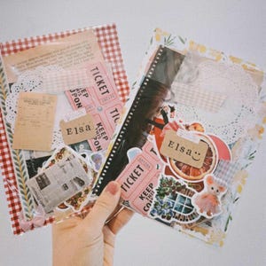 May include: Two clear plastic envelopes filled with colorful paper ephemera, stickers, and decorative elements. The envelopes are decorated with red and white gingham patterns and floral designs. The text "Elsa" appears on both envelopes.
