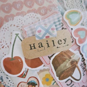 May include: A collection of vintage-style stickers featuring a cherry, a heart with a couple under an umbrella, a flower, a basket, and a label that says "Hailey".