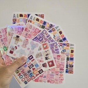 K&ouml;nnte beinhalten: Eine Sammlung von Stickerb&ouml;gen mit bunten Buchstaben, Zahlen und niedlichen Illustrationen. Die Aufkleber zeigen verschiedene Schriftarten und Designs, wobei einige B&ouml;gen holografische Effekte aufweisen. Die Marke "PARTY BLOSSOM" ist auf einem der Stickerb&ouml;gen zu sehen.
