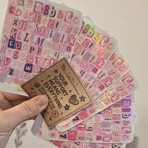 K&ouml;nnte beinhalten: Eine Sammlung rosa Buchstabenaufkleber mit holografischen Akzenten. Die Aufkleber sind f&auml;cherf&ouml;rmig angeordnet und werden von einem kleinen braunen Etikett mit der Aufschrift "YOUR SUPPORT MEANS EVERYTHING" gehalten.