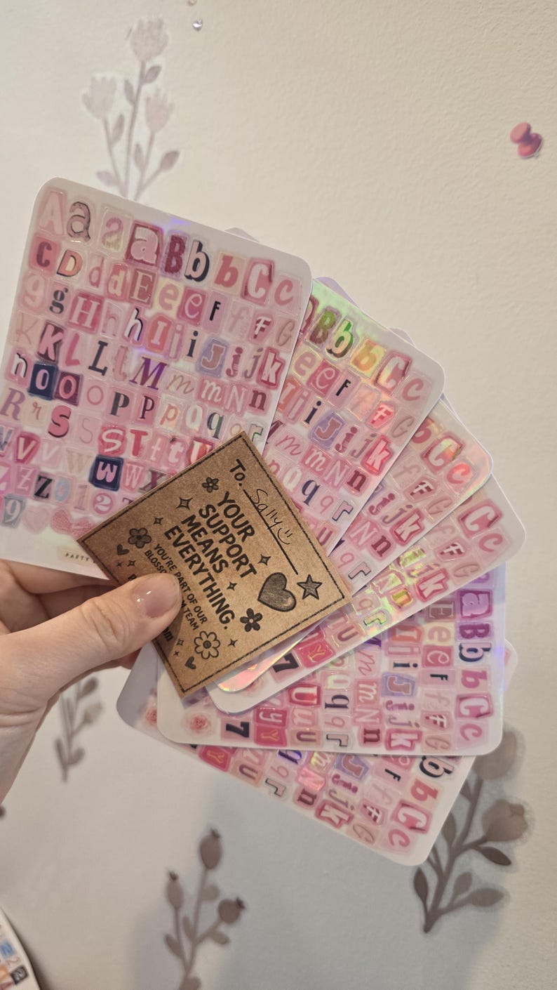 K&ouml;nnte beinhalten: Eine Sammlung rosa Buchstabenaufkleber mit holografischen Akzenten. Die Aufkleber sind f&auml;cherf&ouml;rmig angeordnet und werden von einem kleinen braunen Etikett mit der Aufschrift "YOUR SUPPORT MEANS EVERYTHING" gehalten.