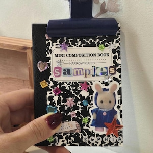May include: A mini composition book with a black and white cow print cover, held by a dark blue binder clip. The cover features the text "MINI COMPOSITION BOOK", "NARROW RULED", "samples", and "SOPHIA", decorated with colorful plastic gems and a rabbit figure.