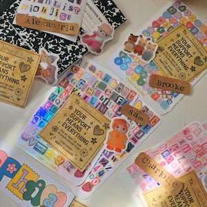 K&ouml;nnte beinhalten: Eine Sammlung dekorativer Aufkleber und Etiketten mit verschiedenen Designs und Texten. Die Aufkleber zeigen Namen wie "Alexandria", "Brooke", "Sam", "Charlotte" und "Sophia". Einige Aufkleber haben ein florales Thema.