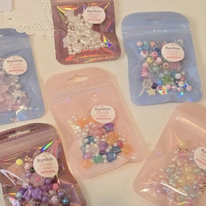 May include: Five resealable pouches containing assorted craft beads in various shapes, sizes, and colors. Each pouch features a circular label with the text Random Beads. The bags are iridescent, light blue, and light pink, holding star, heart, and round beads.