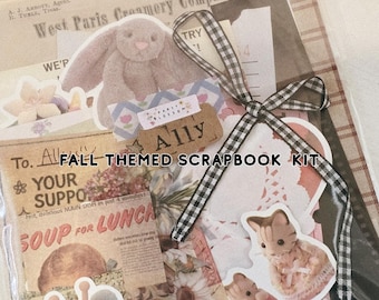 May include: A clear plastic bag containing a scrapbook kit with vintage-style paper ephemera, including a West Paris Creamery Company document, a rabbit sticker, a koala ladybug sticker, and two cat stickers, tied with a black and white gingham ribbon.