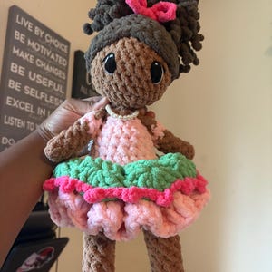 Puede incluir: Muñeca de ganchillo hecha a mano con piel morena, cabello castaño oscuro y un lazo rosa. La muñeca lleva un vestido rosa con una falda con volantes verdes y rosas y un collar de perlas. La muñeca mide unos 30 cm de alto.