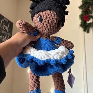Puede incluir: Muñeca de ganchillo hecha a mano con piel morena, cabello negro y un vestido azul y blanco. La muñeca tiene brazos y piernas marrones con detalles azules en los pies y los brazos. La muñeca mide aproximadamente 25 cm de altura.
