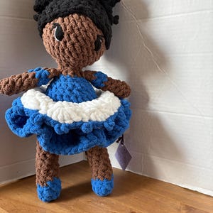Puede incluir: Muñeca de ganchillo hecha a mano con piel morena, cabello negro y un vestido azul y blanco. La muñeca tiene brazos y piernas marrones con zapatos azules. La muñeca está sobre una superficie de madera.
