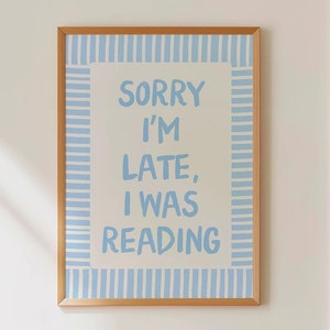 Puede incluir: Un póster enmarcado con el texto "SORRY I'M LATE, I WAS READING" en azul claro sobre un fondo crema. El marco es de color marrón claro. El póster está colgado en una pared blanca sobre una mesa de madera con un libro.