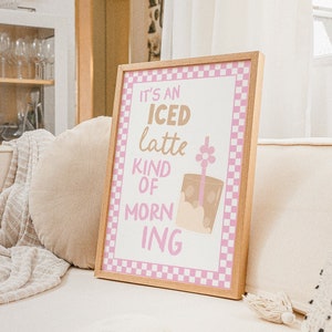 May include: A framed print with a pink and white checkered border. The print features a brown iced latte with a straw and a pink flower on top. The text on the print reads "It's an iced latte kind of morning".