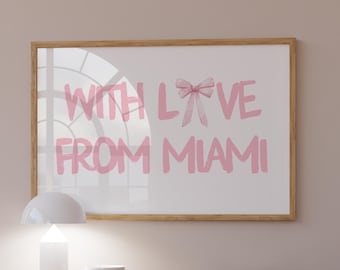 May include: A wooden dresser with three drawers and two doors. The dresser has a light wood finish and is sitting on a light brown wood floor. A white lamp and a white bowl are on top of the dresser. A framed print with the text "With Love From Miami" in pink lettering is hanging on the wall behind the dresser.