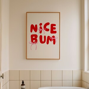 Könnte beinhalten: Ein weißes Badezimmer mit einer weißen Badewanne, einem Holzhocker und einem gerahmten Druck an der Wand, auf dem in roten Buchstaben mit rosa Schleifen "NICE BUM" steht.