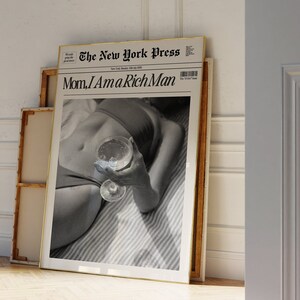 May include: A black and white photo of a person holding a glass of wine. The photo is framed and has the headline "Mom, I Am a Rich Man" from "The New York Press" printed above the image.