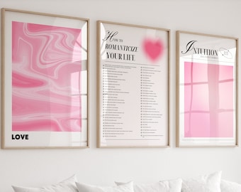 Könnte beinhalten: Drei rosa und weiße gerahmte Poster mit den Worten "LOVE", "Wie man sein Leben romantisiert" und "INTUITION" an einer weißen Wand über einem weißen Sofa mit grünen Kissen und einem Holz-Couchtisch mit einer Vase mit rosa Blumen.
