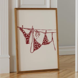 Könnte beinhalten: Gerahmter Kunstdruck mit einem rot-weiß gepunkteten Bikini, der an einer Wäscheleine hängt. Das Kunstwerk ist eine minimalistische Illustration mit hellbeigem Hintergrund und Holzrahmen. Das Bild lehnt an einer weißen Tür.