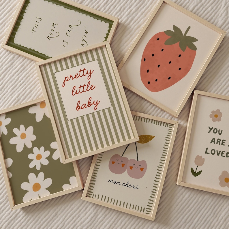 May include: A collection of framed art prints with various designs. The prints include a strawberry, cherries, daisies, and text that reads "pretty little baby", "this room is for playin'", and "you are so loved". The frames are light wood.