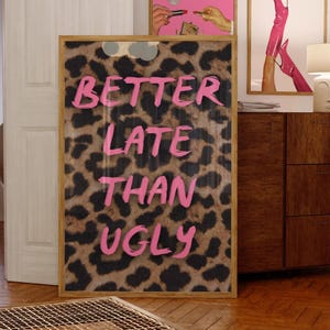 May include: A framed artwork with a leopard print background and the phrase "BETTER LATE THAN UGLY" in pink lettering. The artwork is in a wooden frame and leans against a wall. Other artwork is visible in the background.