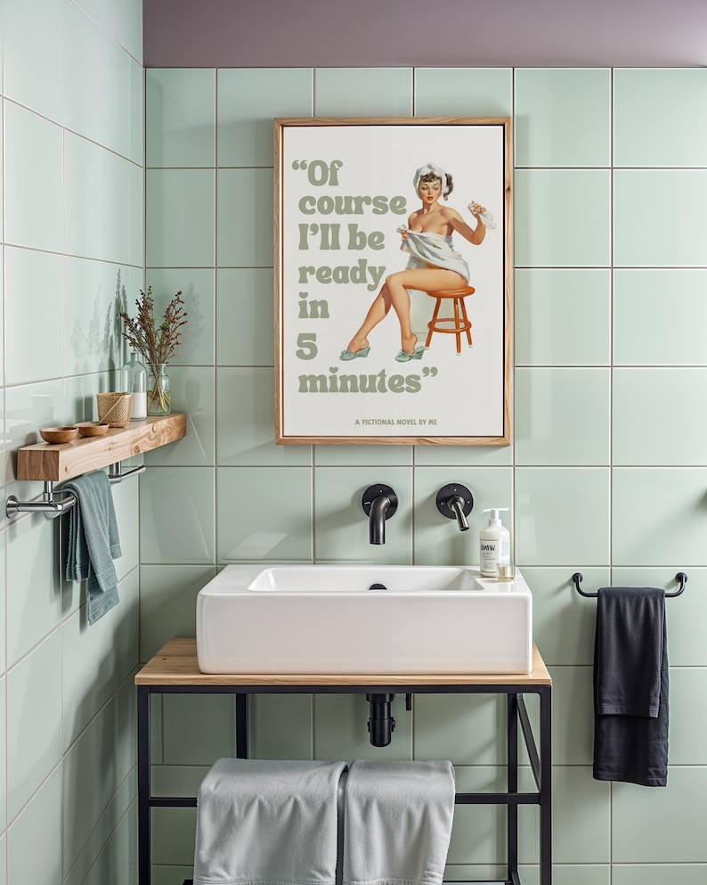 May include: A framed print with a vintage illustration of a woman in a towel, with the text "Of course I'll be ready in 5 minutes." The print is hanging in a bathroom with green tile walls and a white sink.