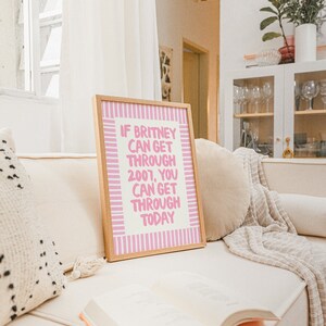 K&ouml;nnte beinhalten: Ein gerahmtes Poster mit rosa und wei&szlig;en Streifen und dem Text "Wenn Britney 2007 &uuml;berstehen konnte, kannst du auch heute durchkommen". Das Poster liegt auf einem wei&szlig;en Sofa mit einem Buch und einer Wolldecke.