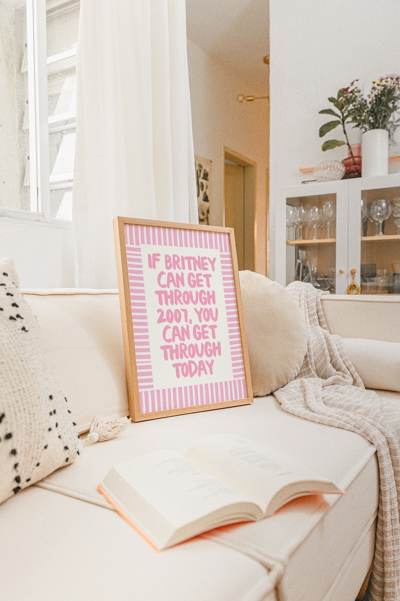 K&ouml;nnte beinhalten: Ein gerahmtes Poster mit rosa und wei&szlig;en Streifen und dem Text "Wenn Britney 2007 &uuml;berstehen konnte, kannst du auch heute durchkommen". Das Poster liegt auf einem wei&szlig;en Sofa mit einem Buch und einer Wolldecke.