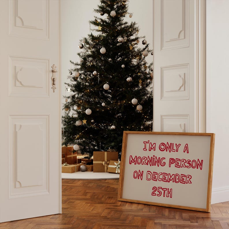 May include: A white framed sign with the text "I'm only a morning person on December 25th" written in red. The sign is leaning against a Christmas tree decorated with white ornaments and lights.