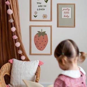 May include: Three framed art prints with botanical and fruit illustrations. One print says "YOU ARE SO LOVED", another says "pretty little baby", and the third features a strawberry. A wicker chair with a white pillow and pink tassels is in the foreground.