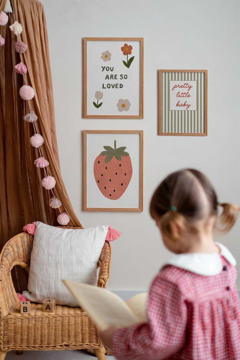 May include: Three framed art prints with botanical and fruit illustrations. One print says "YOU ARE SO LOVED", another says "pretty little baby", and the third features a strawberry. A wicker chair with a white pillow and pink tassels is in the foreground.
