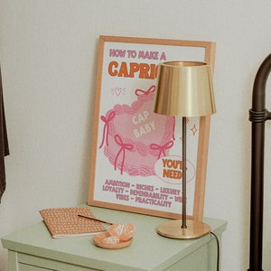 May include: A framed poster with the text "How to Make a Capri" and a pink illustration of a pillow with the text "Cap Baby" and "You'll Need". The poster is on a light green nightstand with a gold table lamp and a small plate of orange slices.
