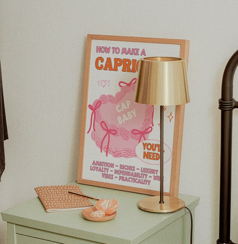 May include: A framed poster with the text "How to Make a Capri" and a pink illustration of a pillow with the text "Cap Baby" and "You'll Need". The poster is on a light green nightstand with a gold table lamp and a small plate of orange slices.