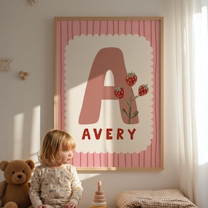 Op de afbeelding: Een ingelijste roze en witte wandkunstprint met de letter "A" in een zachte roze tint, versierd met aardbeienillustraties. De naam "AVERY" is in een vet, rood lettertype gedrukt. Het kunstwerk is geplaatst tegen een roze gestreepte achtergrond.