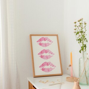 May include: A framed print with three pink watercolor lips on a white background. The print is sitting on a wooden dresser with a vase of white flowers, a candle, and a notebook.