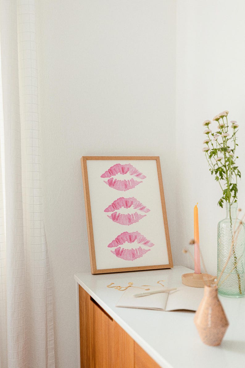 May include: A framed print with three pink watercolor lips on a white background. The print is sitting on a wooden dresser with a vase of white flowers, a candle, and a notebook.