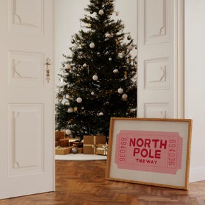 May include: A framed print of a pink ticket with the text "North Pole This Way" and the numbers "694036" on either side. The print is leaning against a white wall next to a decorated Christmas tree.