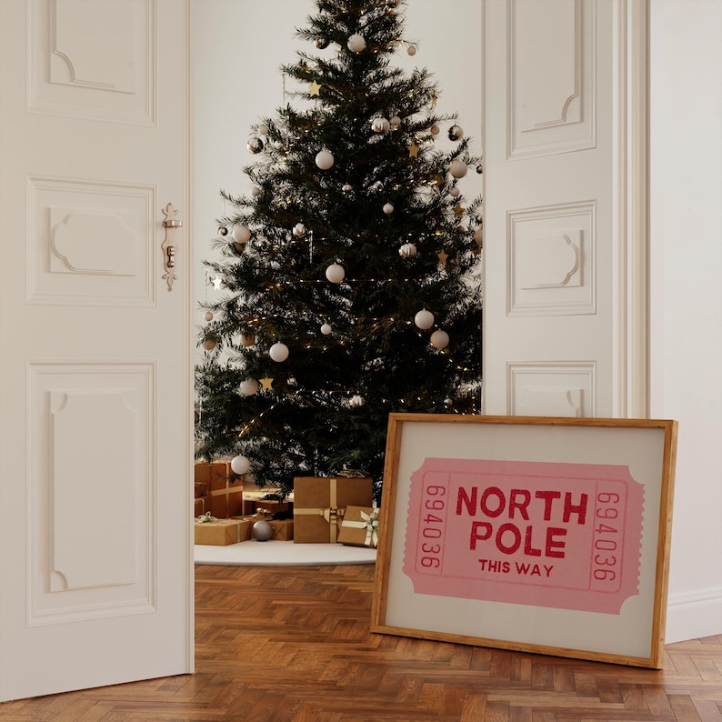 May include: A framed print of a pink ticket with the text "North Pole This Way" and the numbers "694036" on either side. The print is leaning against a white wall next to a decorated Christmas tree.