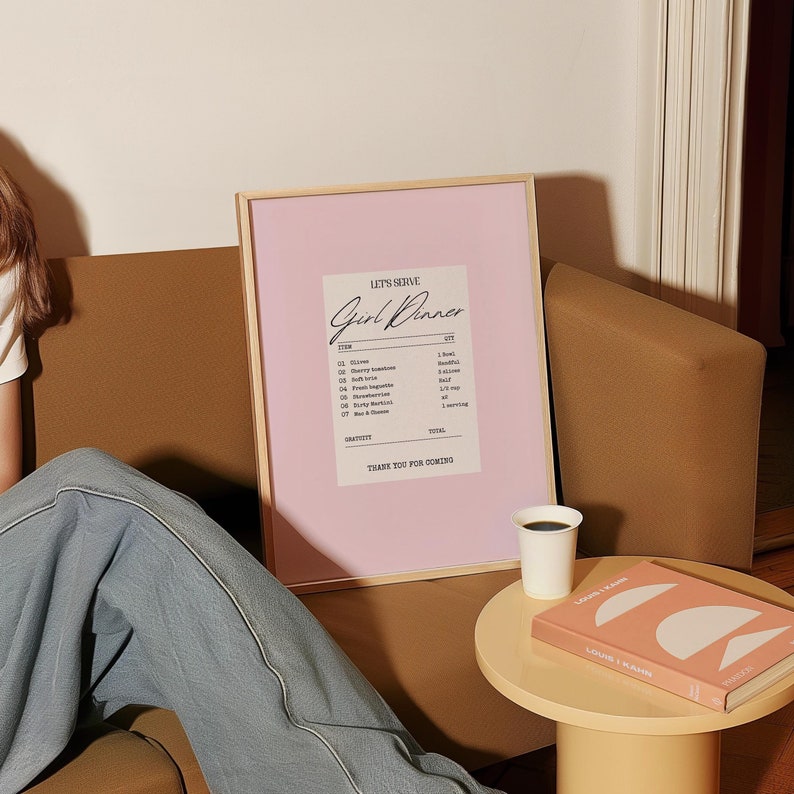 K&ouml;nnte beinhalten: Ein gerahmter Druck mit rosa Hintergrund und einem wei&szlig;en Design im Stil einer Quittung, auf dem "Let's Serve Girl Dinner" steht. Der Druck listet Artikel wie "Oliven", "Cherrytomaten", "Brie", "frisches Baguette", "natives Oliven&ouml;l extra", "Beeren und K&auml;se" und "Fleisch und K&auml;se" auf.