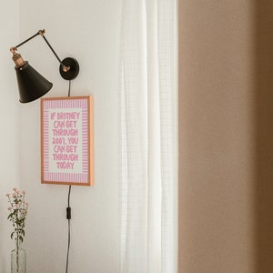 K&ouml;nnte beinhalten: Ein gerahmter rosa-wei&szlig;er Motivationszitat-Druck mit dem Text "Wenn Britney 2007 &uuml;berstehen kann, kannst du es auch heute." h&auml;ngt an einer wei&szlig;en Wand neben einer schwarzen Wandlampe.