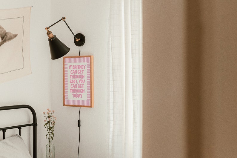 K&ouml;nnte beinhalten: Ein gerahmter rosa-wei&szlig;er Motivationszitat-Druck mit dem Text "Wenn Britney 2007 &uuml;berstehen kann, kannst du es auch heute." h&auml;ngt an einer wei&szlig;en Wand neben einer schwarzen Wandlampe.