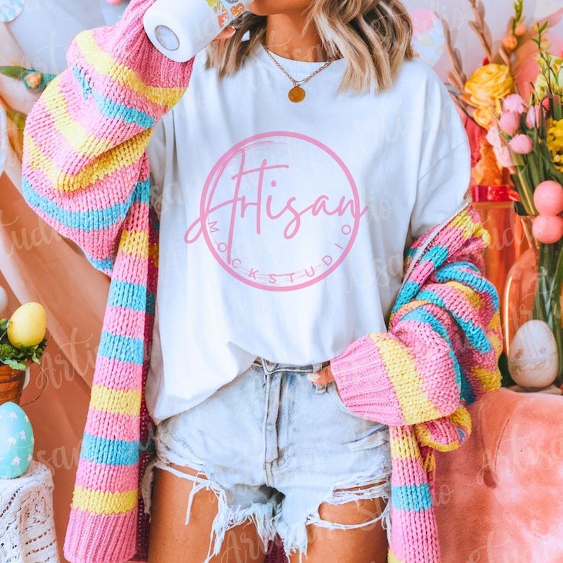 K&ouml;nnte beinhalten: Ein wei&szlig;es T-Shirt mit einem rosa Kreis und dem Text "Artisan Mock Studio" in wei&szlig;er Schrift. Das Shirt wird von einer Person mit einem rosa, gelben und blauen gestreiften Cardigan getragen.
