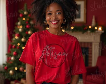 May include: A woman wearing a red t-shirt with a white circle and the word "Artisan" in pink lettering. She is standing in front of a Christmas tree and a fireplace.