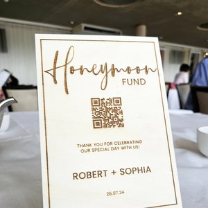 May include: A wooden sign with the text "Honeymoon Fund" and a QR code. The sign also includes the names "Robert + Sophia" and the date "26.07.24".