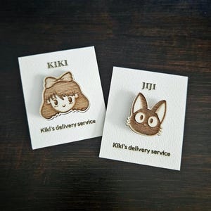 May include: Two wooden pins featuring characters from the animated film Kiki's Delivery Service. The first pin is of Kiki, a young witch with brown hair and a red bow. The second pin is of Jiji, Kiki's black cat companion. Both pins have the text "Kiki's delivery service" on the card.