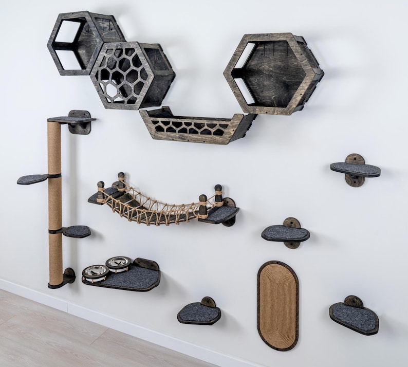 May include: A collection of cat furniture mounted on a white wall. The set includes hexagon-shaped shelves, a bridge, a scratching post, and various platforms. The pieces are made of wood and gray felt, with a natural rope bridge.