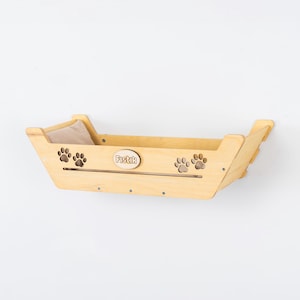 May include: A light-colored wooden cat bed with a beige cushion inside. The bed is mounted on a wall and features paw print cutouts and the name "Fistik" engraved on the front.