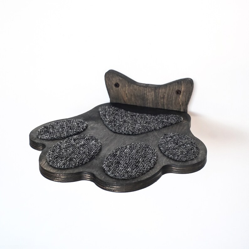 May include: A dark gray wooden cat paw-shaped wall shelf with five gray carpeted pads. The shelf is designed to be mounted on a wall and provides a comfortable spot for a cat to rest or play. The wood has a natural grain.